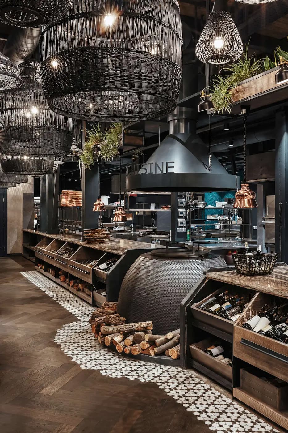 Open kitchen and dining counter beneath a sculptural black hood, warm lighting and suspended greenery.
