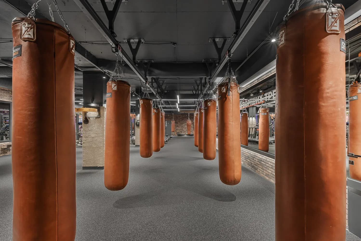 Row of orange heavy bags suspended beneath exposed black ceiling services.